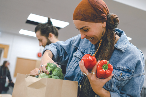A smiling woman wearing a headscarf and denim jacket holds two red bell peppers and places broccoli into a paper bag. Another person is in the background, also packing groceries.