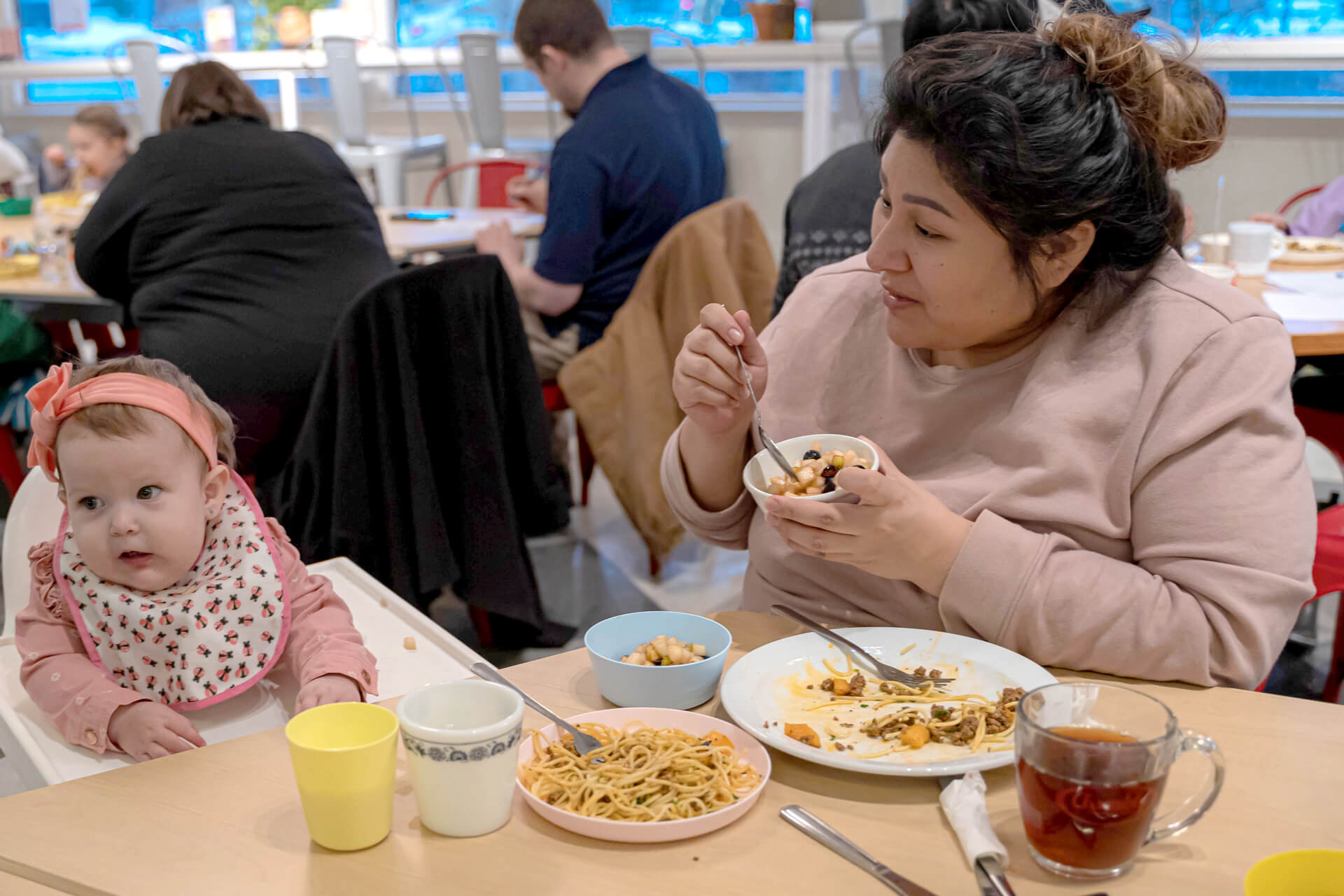 A woman eats at a table with a baby in a high chair. The table has plates of food, cups, and a glass of tea. The baby wears a bib and headband, looking toward the camera in a busy restaurant setting.