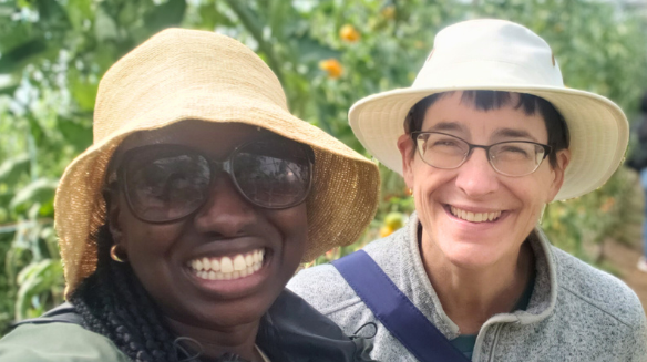 Two women wearing wide-brimmed hats and glasses smile for a selfie in an outdoor garden with green plants and yellow flowers in the background.